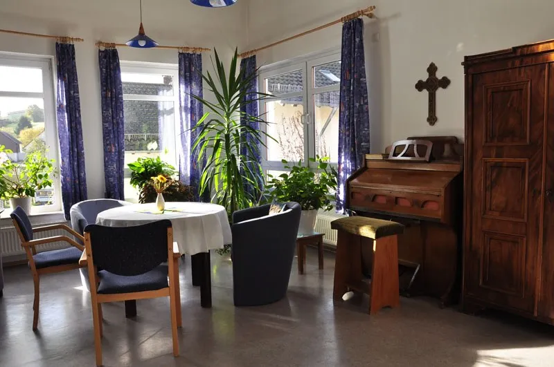 Heller Raum mit Sesseln, Pflanzen und einem Kreuz an der Wand